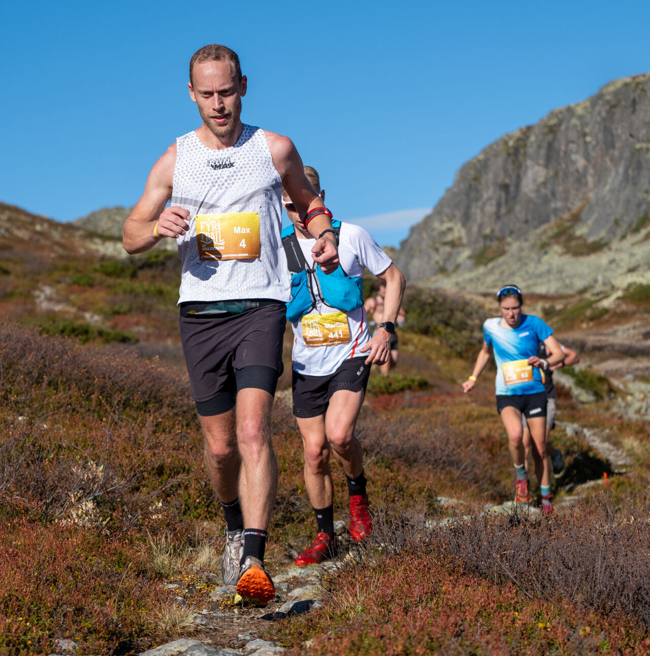 Fýri Trail photo Magnus Grönberg web 09-24-23 Trail runners wearing race bibs at Fýri Trail by Salomon along a rocky mountain path beneath a clear blue sky