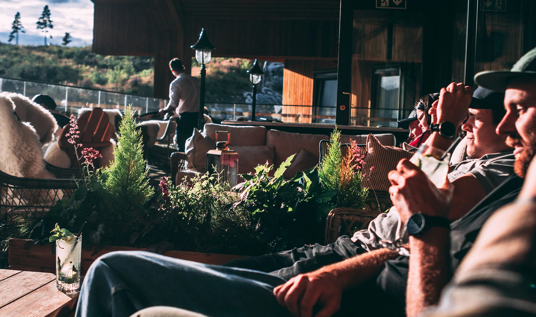 People relax in a sunny outdoor lounge surrounded by potted plants and mountain scenery