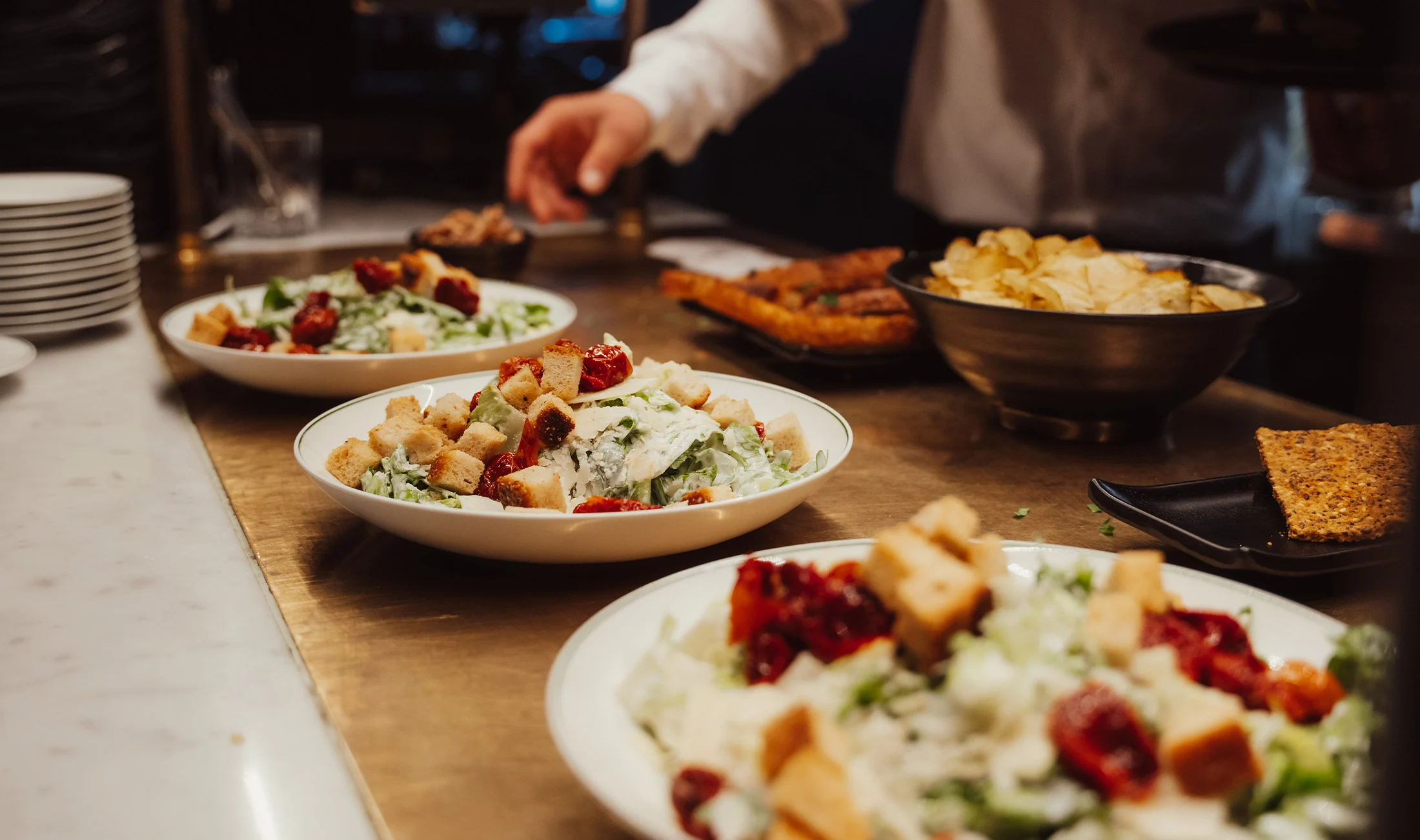 Salad bowls with greens, croutons, sun-dried tomatoes, bread, and chips arranged on a restaurant counter