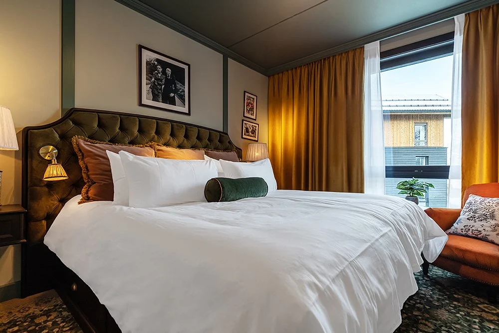 Standard_Double_Fyri_Resort Bedroom with a tufted headboard, white bedding, gold curtains, framed art, and a window overlooking neighboring buildings